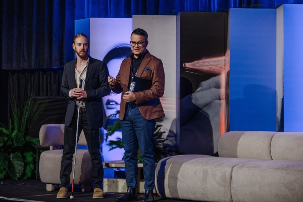 Two presenters stand on stage at The Outlook in front of bold backdrop panels, preparing to speak with microphones in hand.