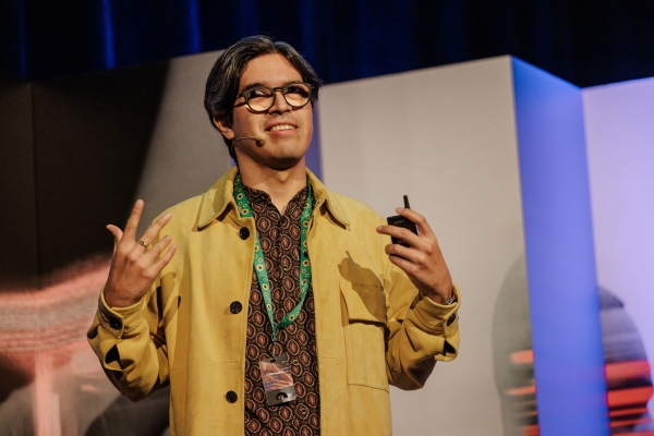 Speaker wearing a yellow jacket and patterned shirt presents confidently on stage at The Outlook, smiling and gesturing expressively.