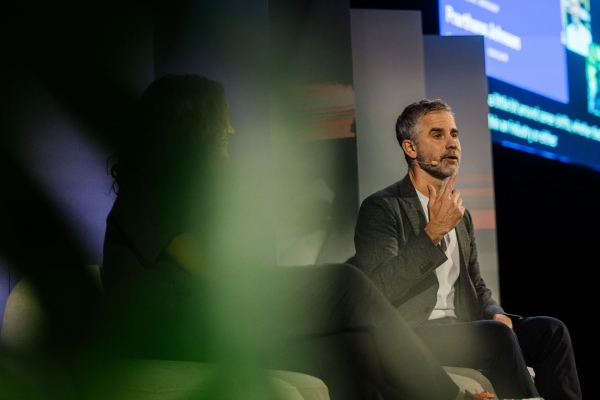 Prathana Johnson and Alex Burke in a fireside chat on the Design Outlook 2025 stage, partially obscured by greenery in the foreground.
