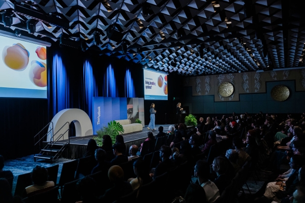 Julia Suh presenting on stage at Design Outlook 2025, speaking to a full audience with slides asking, “Are we designing living, breathing systems?"