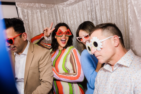 Sarah D’Sylva and friends in playful sunglasses at TO25, posing in a photo booth with Sarah throwing a peace sign and smiling brightly.