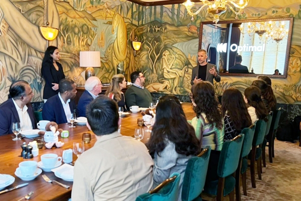 A group of people sit around a long dining table listening to a speaker, with a mural backdrop and chandelier lighting in a formal meeting setting.