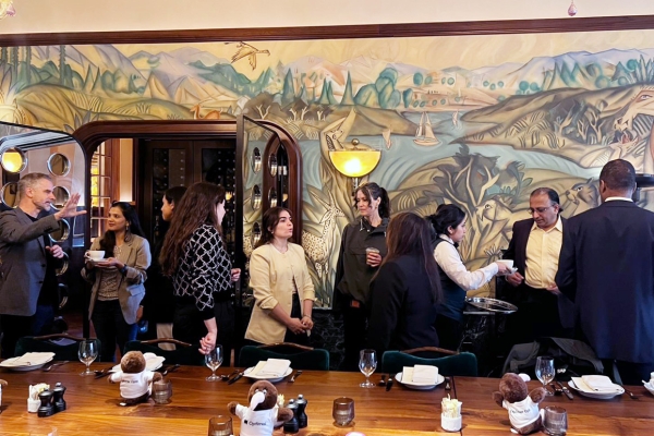 People gather and converse in a warmly lit dining room with a mural backdrop, set table in foreground with plush toys placed on plates.