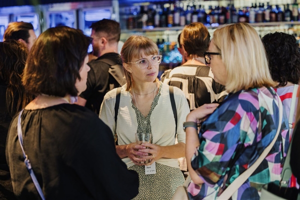 A woman holding a drink listens attentively while conversing with two others at a crowded networking event near a bar.