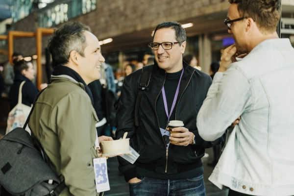 Three people chat and smile while holding coffee and food containers in a bright, open event space with others mingling in the background.