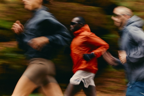 A dynamic, motion-blurred image of three runners moving through a forest trail. One runner in the center wears a bright orange jacket, standing out against the earthy tones of the background. The blurred motion emphasizes speed, energy, and the natural setting of the run.