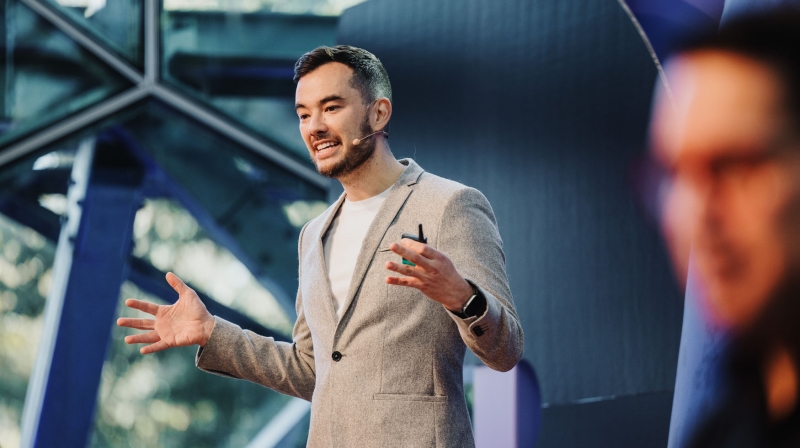 Presenter, Jonathan Lau, on stage at Design Outlook 2023, speaking with animated gestures while wearing a grey blazer and headset mic.