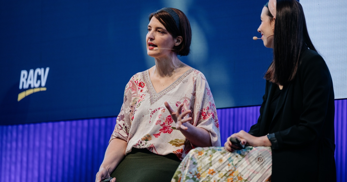 Two women sitting on stage in conversation, both wearing headset microphones. One woman, wearing a floral blouse and green skirt, is speaking while gesturing with her hand. The other woman, wearing a black blazer and a floral skirt, is listening. The RACV logo is visible on a large screen in the background.