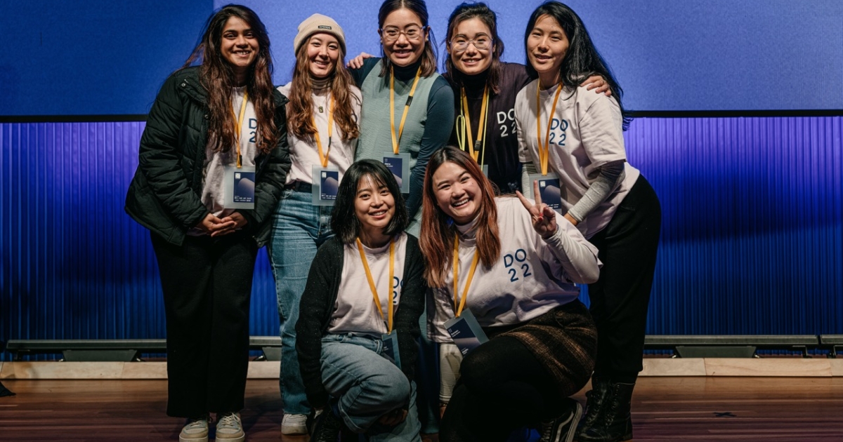 A group of seven volunteers at the DO 2022 conference pose together in front of a blue backdrop on a stage. They are smiling warmly, with a few of them making peace signs. Most are wearing white DO 22 t-shirts, and all have conference badges and lanyards. Their expressions reflect a sense of camaraderie and enthusiasm, capturing the welcoming atmosphere of the event.