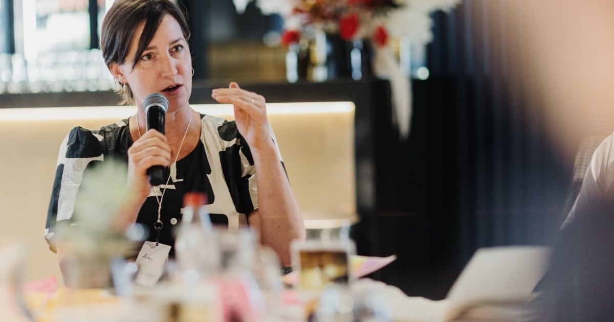 Amy Sheppard speaking into a microphone during a discussion at a leadership event. She is wearing a patterned black and white dress and gesturing with one hand. The background includes soft lighting and a floral arrangement, adding a professional and warm atmosphere to the setting.