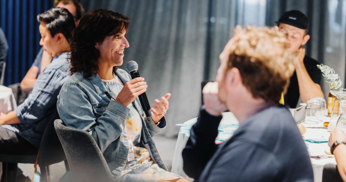 A woman seated at a table speaking into a microphone during a leadership event. She is smiling and wearing a denim jacket over a colorful outfit. Others at the table listen attentively, while the softly lit room and curtains in the background create a focused yet relaxed atmosphere.