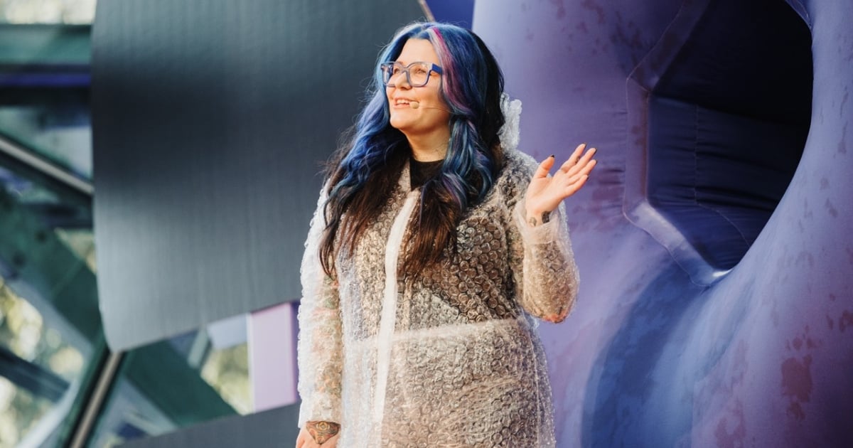 Chi Ryan presenting at DO23, wearing a transparent bubble wrap coat over dark clothing, with blue and purple-streaked hair and clear-rimmed glasses. Chi stands on stage with a colorful, abstract background, smiling and gesturing with one hand raised, engaging the audience.