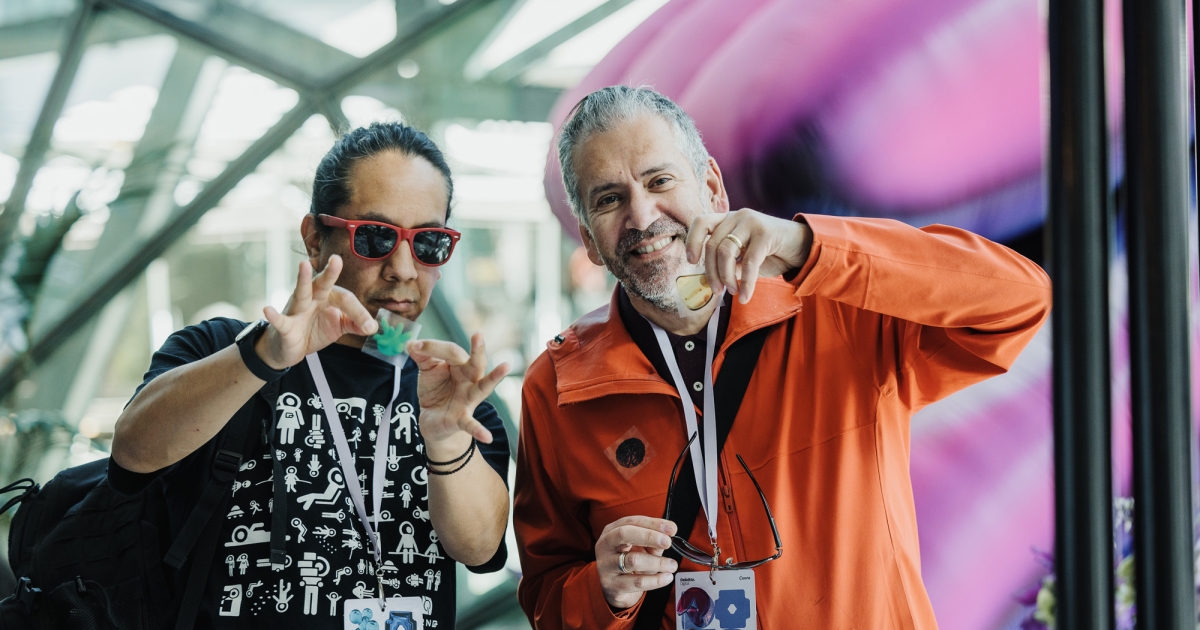 Two individuals at a Design Outlook event interacting with colorful materials at a display table. The person on the left is wearing red sunglasses, a black graphic t-shirt, and holding a green object, while the person on the right, dressed in an orange jacket, smiles while holding a yellow object. The vibrant background features abstract shapes and a modern, light-filled setting.