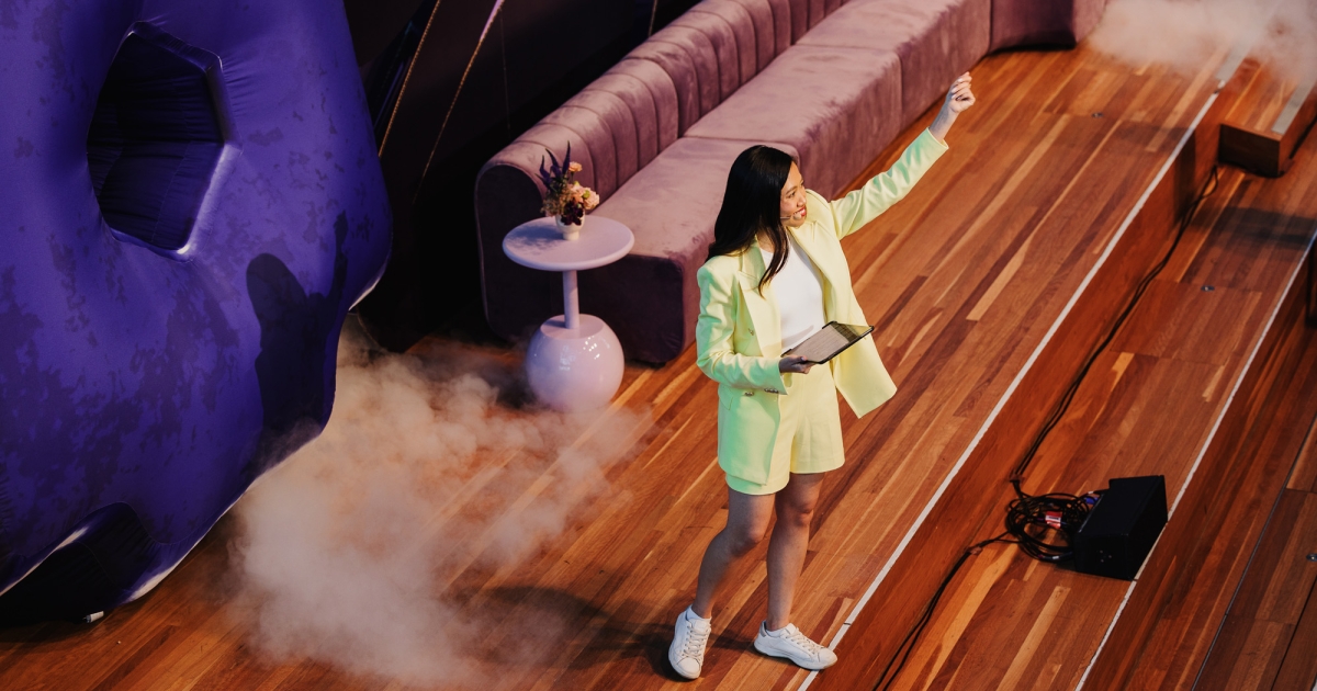 Berlin Liew presenting at a Design Outlook event, captured from an elevated angle. She is wearing a stylish light yellow blazer and shorts with white sneakers, holding a tablet in one hand while gesturing confidently with the other. The stage features a purple sofa, decorative side tables, and atmospheric smoke effects, with wooden floors and a modern, dynamic setup enhancing the scene.