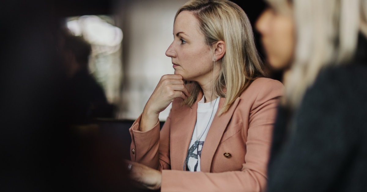 A woman with blonde hair, wearing a light peach blazer over a white top, sits thoughtfully with her hand resting on her chin. She appears focused, looking towards the right side of the frame. The background is softly blurred, emphasizing her contemplative expression during what seems to be a conference or meeting setting.