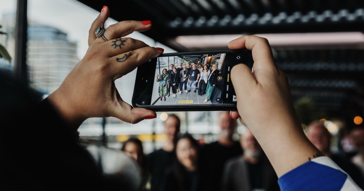 A close-up of a person’s hands holding a smartphone horizontally, capturing a group photo of people posing together in an outdoor covered area. The person’s hands feature bright red nail polish and tattoos of a sun and crescent moon on their fingers. The smartphone screen shows a clear image of the group, framed by blurred background elements in the real scene.