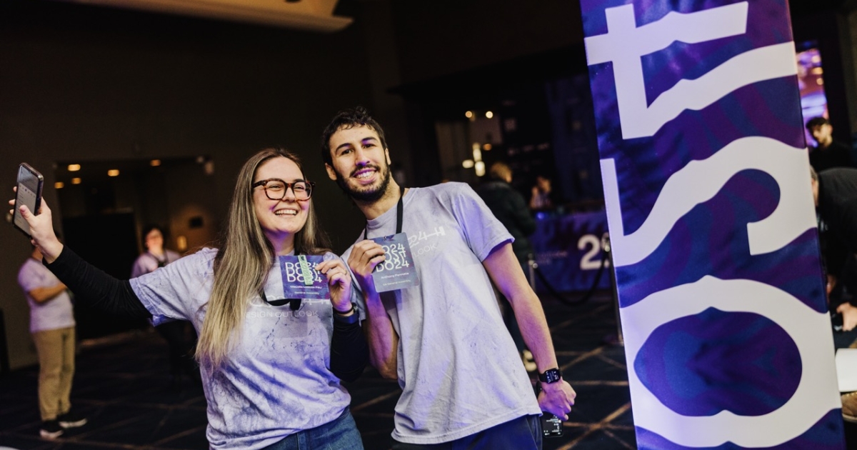 Two enthusiastic volunteers at the DO 2024 conference pose together, smiling and holding their conference badges. Both are wearing event t-shirts and lanyards. One of the volunteers holds up a phone for a selfie. They stand near a tall, colorful banner featuring abstract patterns in purple and white with the DO24 branding