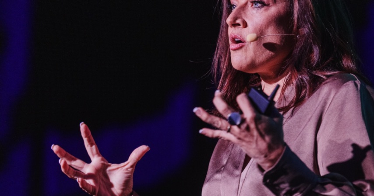 Alison Page presenting at DO24, captured mid-speech with expressive hand gestures. She is wearing a silky, taupe-colored blouse and a leopard print belt, standing against a dark background with dramatic lighting that highlights her focused expression.