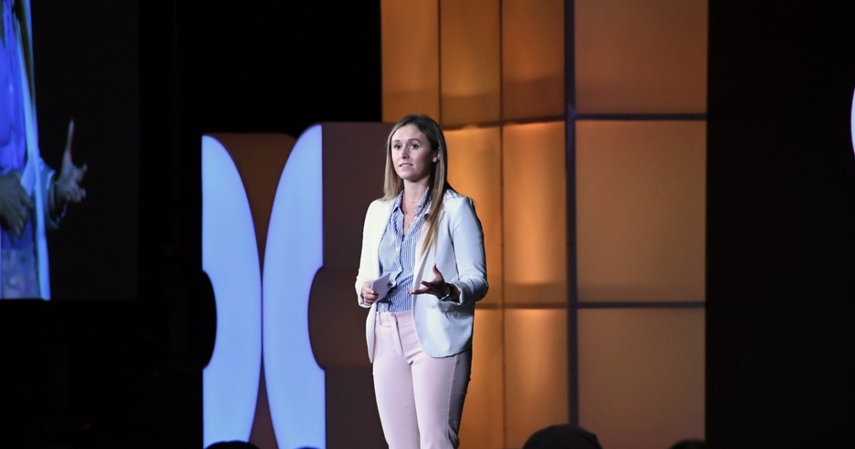 A woman in a white blazer and light trousers speaks on stage under warm lighting, addressing an audience with a large event screen behind her.