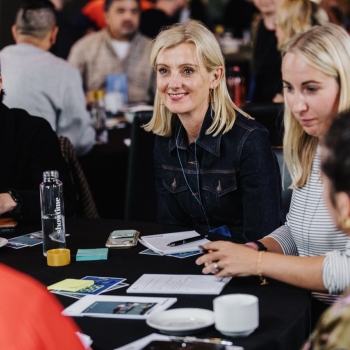 A group of people sitting at a table during a conference, with a blonde woman in a denim jacket smiling in the centre