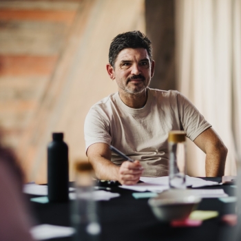 A man with short dark hair and a beard, wearing a light-coloured T-shirt, sitting at a table during the Design Leadership event, holding a pen and looking thoughtful