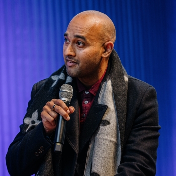 Abi Ayer peaking into a microphone. He is wearing a dark coat and a patterned scarf, with a purple backdrop behind him