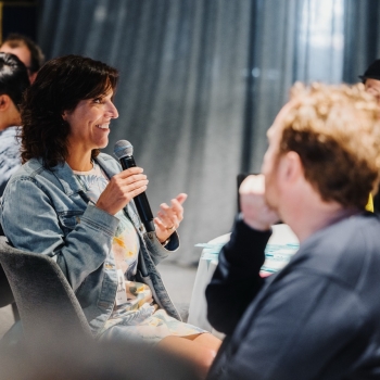 A woman seated at a table speaking into a microphone during a leadership event. She is smiling and wearing a denim jacket over a colorful outfit. Others at the table listen attentively, while the softly lit room and curtains in the background create a focused yet relaxed atmosphere.