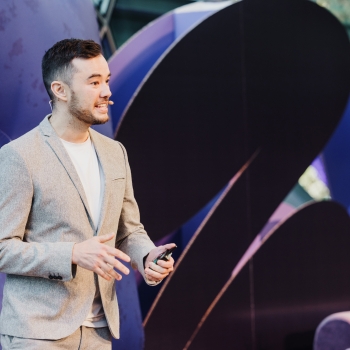 Jon speaking at a Design Outlook event, dressed in a light gray suit and a white shirt. He holds a small clicker in his hand and gestures confidently while addressing the audience. The background features abstract purple and blue shapes, creating a vibrant and modern atmosphere.