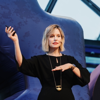 MC Monsalve presenting at a Design Outlook event, gesturing with one hand raised while holding a clicker in the other. She is wearing a black outfit with a statement gold necklace and a headset microphone, with a confident and poised expression. The backdrop features abstract blue shapes and modern architectural elements, enhancing the event’s contemporary ambiance.