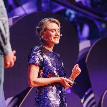 Dr. Barbara Doran presenting at a Design Outlook event, smiling confidently while speaking into a headset microphone. She is wearing a shimmering purple sequined dress, holding a clicker in one hand. The background features modern stage design elements illuminated with purple lighting, creating a vibrant and engaging atmosphere.