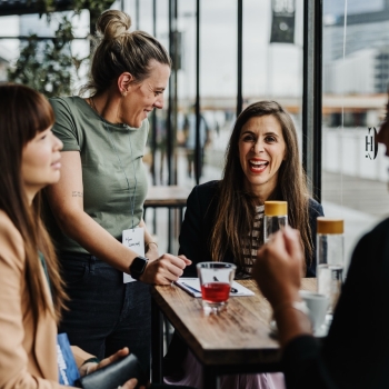 A lively moment in a café-like setting with four women chatting around a wooden table near large windows. One stands smiling in a green t-shirt, while another seated in a striped top and blazer laughs warmly. The setting feels modern and friendly, with an urban view outside.