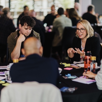 A group of professionals engaged in a lively discussion around a round table during a leadership event. A woman in glasses and a black blazer is speaking energetically, while others listen attentively. The table is covered with notes, notebooks, and water bottles, with a collaborative atmosphere reflected in the background of other groups in conversation