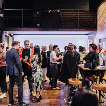 Guests networking during drinks after an event, engaging in lively conversations in a stylish indoor venue. The space features warm wooden accents, high-top tables with stools, and a softly lit backdrop, creating a relaxed and collaborative atmosphere to conclude the event.