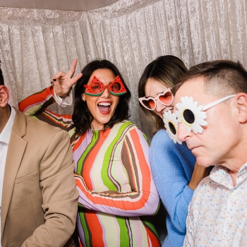 Sarah D’Sylva and friends in playful sunglasses at TO25, posing in a photo booth with Sarah throwing a peace sign and smiling brightly.