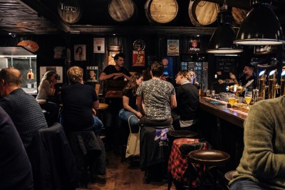 A lively, rustic pub with people chatting at tables and the bar. Warm lighting, vintage decor, and full beer glasses create a relaxed vibe.