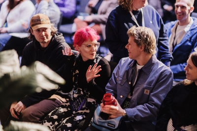 DO24 attendees in animated conversation before a session, with a woman in pink hair gesturing expressively while others listen in the lively audience setting.