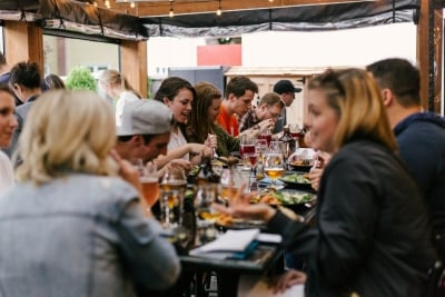 Large group of people gathered at a long outdoor table, enjoying food and drinks together in a lively, social dining setting.