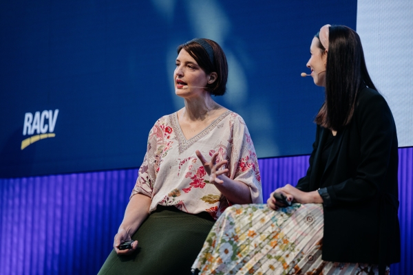 Two women sitting on stage in conversation, both wearing headset microphones. One woman, wearing a floral blouse and green skirt, is speaking while gesturing with her hand. The other woman, wearing a black blazer and a floral skirt, is listening. The RACV logo is visible on a large screen in the background.