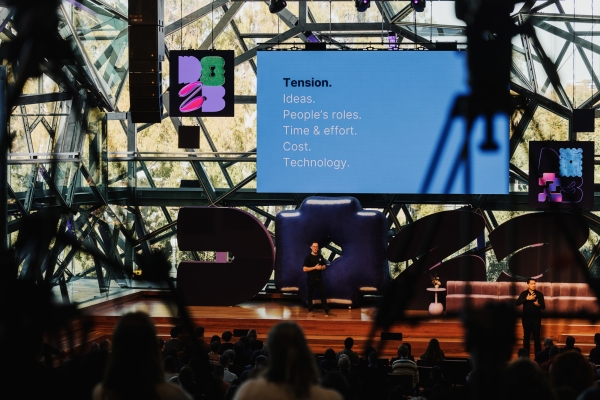 A presenter stands on stage in front of a large screen displaying a slide with the title ‘Tension’ and a list: ‘Ideas, People’s roles, Time & effort, Cost, Technology.’ The stage is set in a large, modern venue with a geometric glass structure and audience members seated in the foreground.