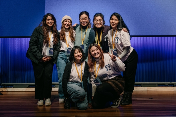 A group of seven volunteers at the DO 2022 conference pose together in front of a blue backdrop on a stage. They are smiling warmly, with a few of them making peace signs. Most are wearing white DO 22 t-shirts, and all have conference badges and lanyards. Their expressions reflect a sense of camaraderie and enthusiasm, capturing the welcoming atmosphere of the event.