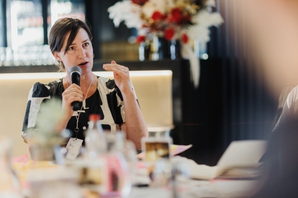 Amy Sheppard speaking into a microphone during a discussion at a leadership event. She is wearing a patterned black and white dress and gesturing with one hand. The background includes soft lighting and a floral arrangement, adding a professional and warm atmosphere to the setting.