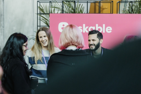 Attendees engaging in conversation at a networking event in front of a pink ‘Askable’ banner. A woman in a striped sweater and a man with a beard are smiling, while others, partially visible from behind, join the discussion. The setup includes plants and a casual, friendly atmosphere.