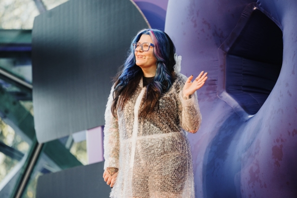 Chi Ryan presenting at DO23, wearing a transparent bubble wrap coat over dark clothing, with blue and purple-streaked hair and clear-rimmed glasses. Chi stands on stage with a colorful, abstract background, smiling and gesturing with one hand raised, engaging the audience.