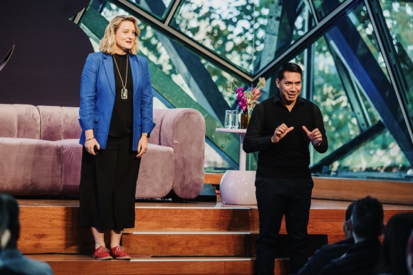 Trudi (left) and Mark (right) addressing the audience on stage at a Design Outlook event. Trudi is wearing a bright blue blazer and black outfit, while Mark, dressed in black, is actively signing as an Auslan interpreter. The stage features a modern setup with a purple sofa and decorative flowers, framed by large glass windows revealing greenery outside.