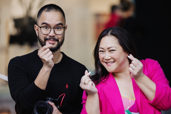 Two individuals posing playfully at a Design Outlook event. On the left, a man with glasses, a beard, and a black sweater holding a camera gestures with his hand. On the right, Jeanette Cheah, wearing a vibrant pink blazer and a colorful top, smiles warmly while mimicking the same gesture. The background is softly blurred, emphasizing their lively and cheerful expressions.