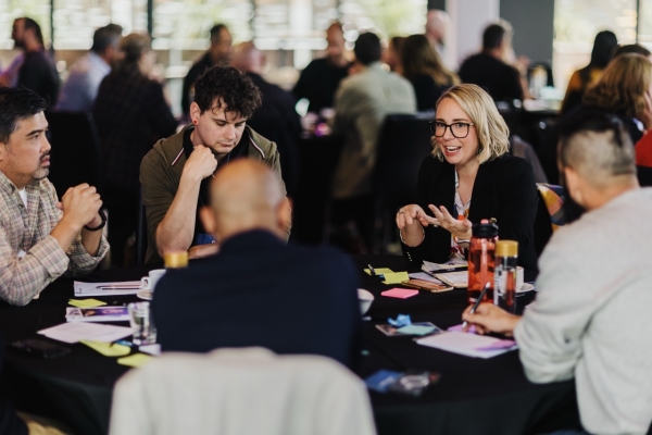 A group of professionals engaged in a lively discussion around a round table during a leadership event. A woman in glasses and a black blazer is speaking energetically, while others listen attentively. The table is covered with notes, notebooks, and water bottles, with a collaborative atmosphere reflected in the background of other groups in conversation