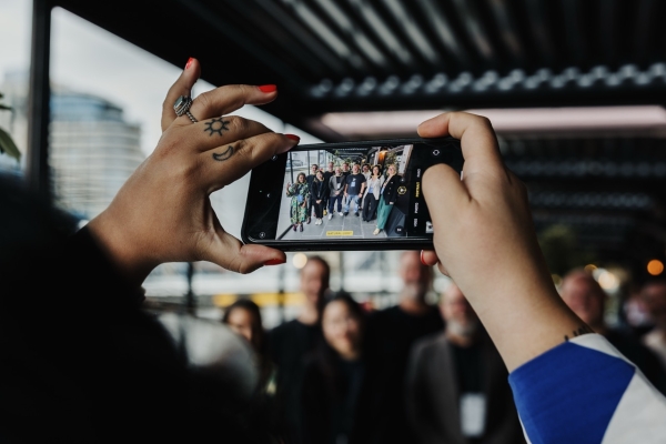 A close-up of a person’s hands holding a smartphone horizontally, capturing a group photo of people posing together in an outdoor covered area. The person’s hands feature bright red nail polish and tattoos of a sun and crescent moon on their fingers. The smartphone screen shows a clear image of the group, framed by blurred background elements in the real scene.