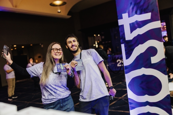 Two enthusiastic volunteers at the DO 2024 conference pose together, smiling and holding their conference badges. Both are wearing event t-shirts and lanyards. One of the volunteers holds up a phone for a selfie. They stand near a tall, colorful banner featuring abstract patterns in purple and white with the DO24 branding