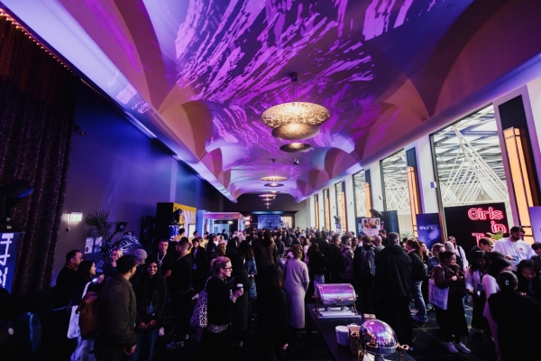 The Sofitel venue for DO24, showcasing a vibrant and bustling networking event. The long, elegant room features dramatic purple lighting projected onto the ceiling, ornate chandeliers, and floor-to-ceiling windows on one side. Attendees fill the space, engaging in conversations and exploring booths such as ‘Girls in Tech,’ with a modern architectural backdrop visible through the windows. The atmosphere is lively and sophisticated, reflecting the energy of the event.