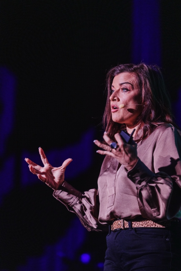 Alison Page presenting at DO24, captured mid-speech with expressive hand gestures. She is wearing a silky, taupe-colored blouse and a leopard print belt, standing against a dark background with dramatic lighting that highlights her focused expression.